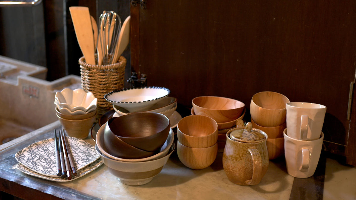 A collection of handmade ceramic and wooden tableware, including bowls, cups, a small teapot, and utensils, arranged on a tabletop against a dark wooden background.