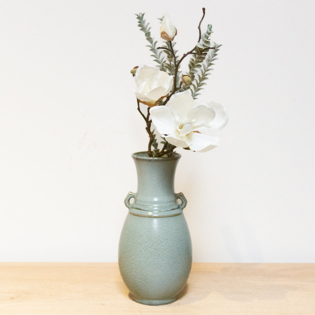 Green ceramic vase with white flowers on a wooden surface and light gray background